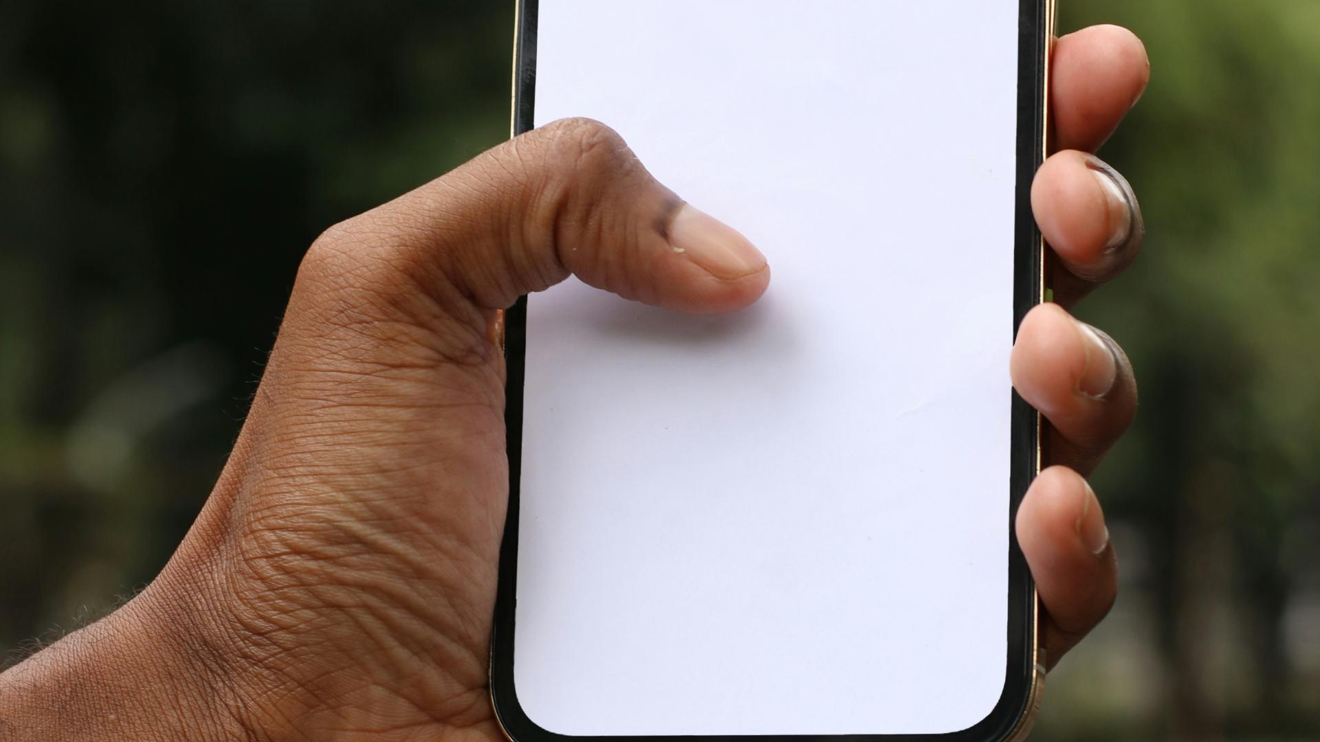 Black hand holding a smartphone with a blank white screen outdoors.