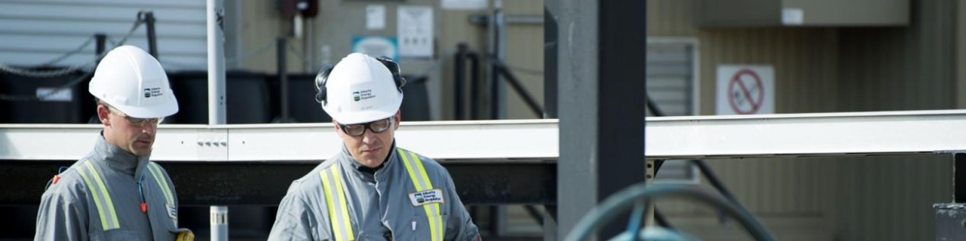 Two caucasian men in their forties with white hard hats on, looking down at a large piece of metal machinery in the foreground, with a white metal beam in the background